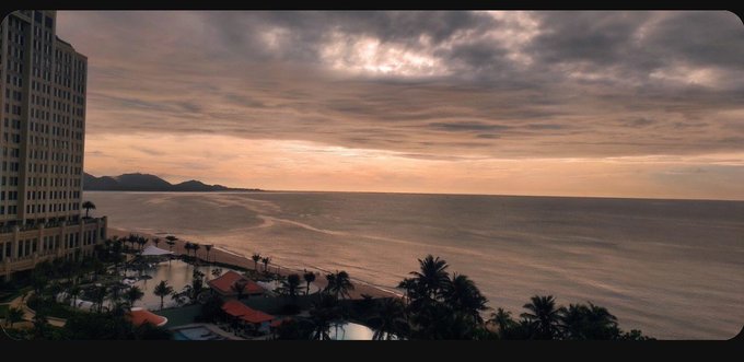 A sunset view over a calm ocean from a high-rise building, with palm trees, a resort pool area, and distant islands visible along the coastline.