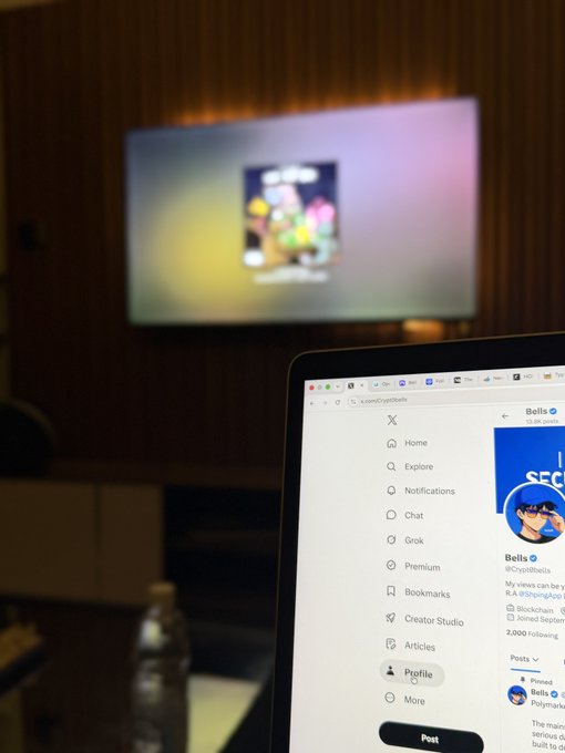 A laptop displays the X (formerly Twitter) sidebar menu with navigation options including "Home," "Explore," "Notifications," "Chat," "Grok," "Premium," "Bookmarks," and a highlighted "Profile" tab, while a blurred TV in the background shows colorful content. The image ties into the "GM!" (Good Morning) post by capturing a casual morning moment of checking social media on a laptop, with the "Profile" tab selected suggesting the user is about to post their morning greeting.