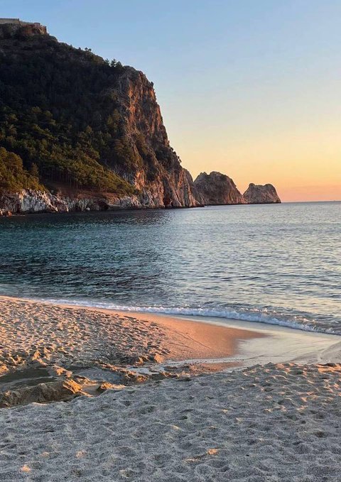 Scenic beach photograph at sunset showing a sandy shore, calm blue water, and a large rocky cliff with vegetation on the left side.