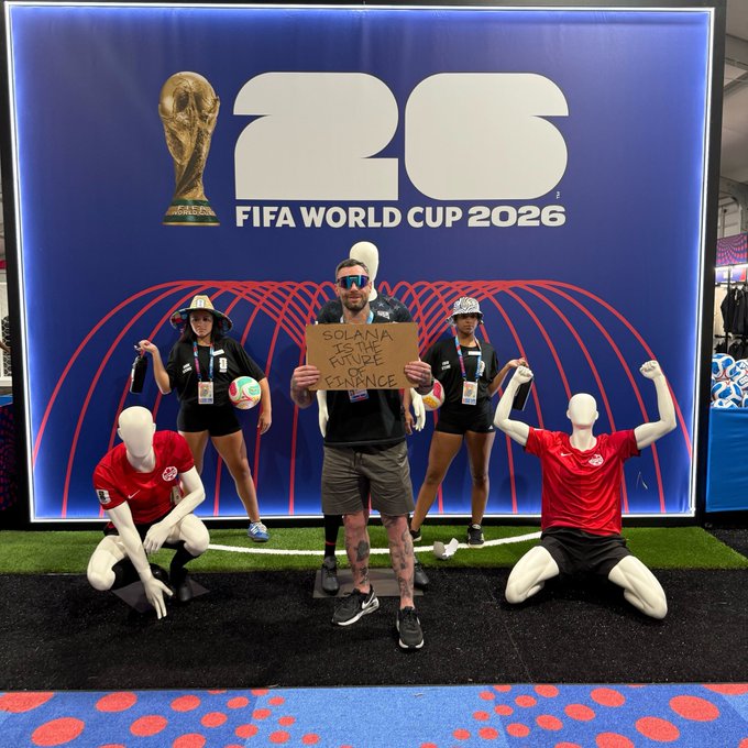The image features a group of people posing for a photo with a mannequin dressed in a red shirt, black shorts, and sunglasses holding up a sign that says "I want to make sure everyone knows @solana is the future of finance at the @FIFAWorldCup @FWC26KansasCity". The mannequin appears to be wearing a red shirt and shorts. There are four people in total posing for the photo - two women and two men, with one woman holding up the sign while the other three pose around her.