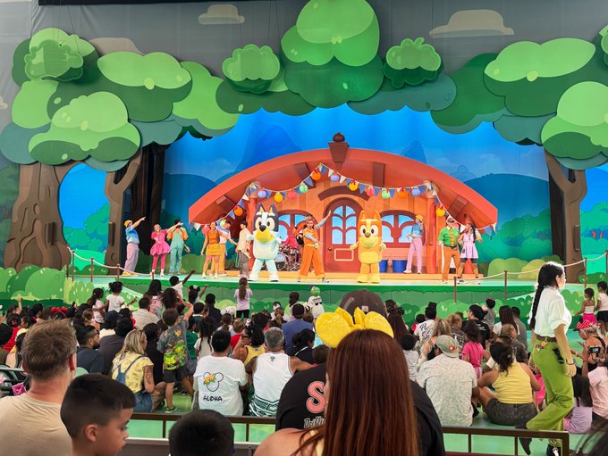 Costumed characters including mascots perform on a brightly decorated stage in front of an audience of children and families.