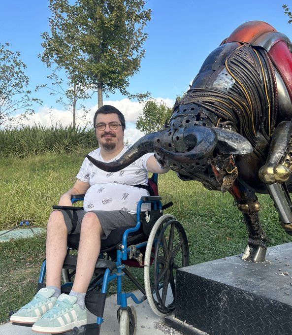 A young man wearing glasses and a white t-shirt with handprints sits in a blue wheelchair outdoors, posing next to a large metallic bull sculpture.