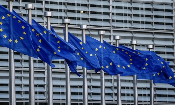 The image features a row of blue flags fluttering in front of a building, with the word "EU" prominently displayed on them. The flags are waving in the wind, creating an atmosphere of unity and patriotism. In the background, there is a building that appears to be a bank or financial institution, as indicated by its presence in the image. The post also mentions several exchanges, including Binance, Coinbase, Kraken, and Crypto.com, which have removed Tether from their platforms due to non-compliance with MiCA rules. This has led to a shift in focus towards more compliant stablecoins like $USDC, which is now considered the leading stablecoin in the EU.