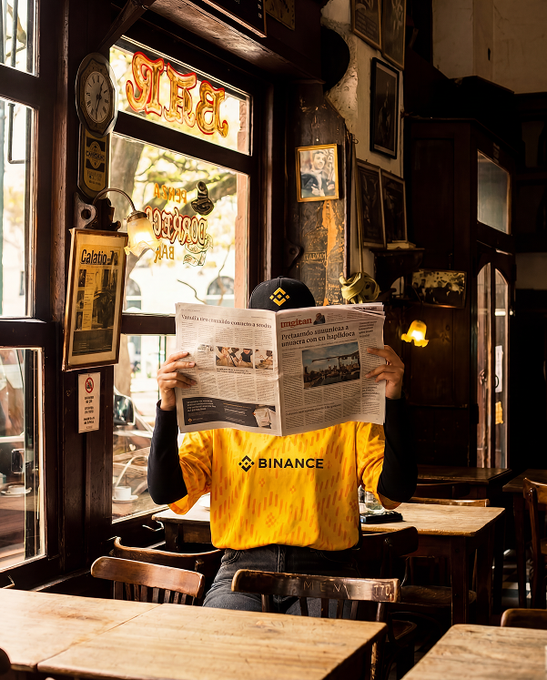 A person wearing a yellow Binance t-shirt and black cap sits in a rustic café/bar reading a Spanish-language newspaper, with vintage decor, framed portraits, and a window visible in the background.