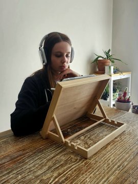 A person wearing headphones sits at a wooden table, looking at a wooden shadow box frame displaying a preserved thorny branch.