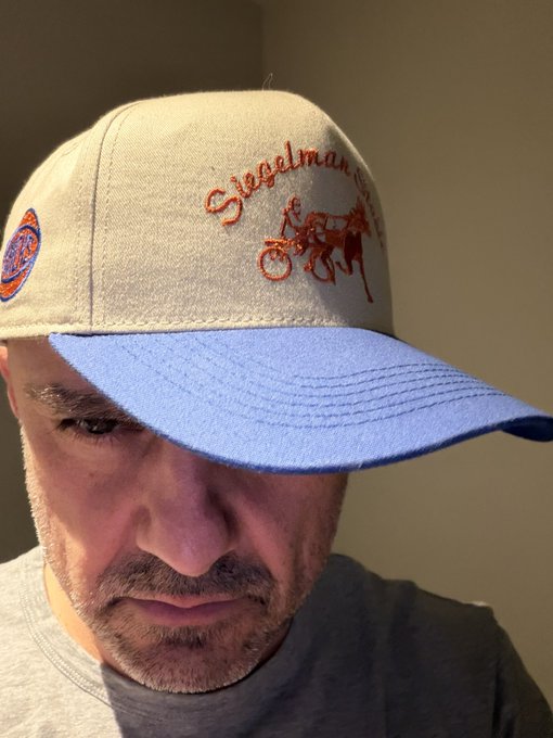 The image features a man wearing a baseball cap with a logo that reads "Siegelsman". He has a serious expression as he looks down at his hat, possibly contemplating something or simply enjoying the moment. The background of the photo appears to be a wall, and there is also an orange object in the scene, which could potentially be related to the Siegelsman logo on the cap.