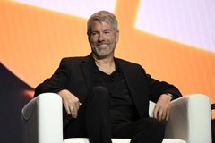 A bearded man in a black suit sits relaxed in a white chair against a bright orange backdrop, consistent with the post's reference to Michael Saylor, the prominent Bitcoin advocate and MicroStrategy executive, appearing at a public event or conference.