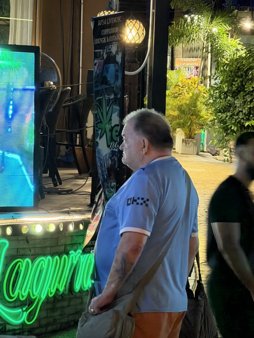 A man in a blue shirt stands at night near a neon 'Laguna' sign and a screen displaying graphs, holding a bag.
