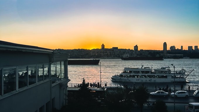 A sunset over a city skyline with a body of water in the foreground, featuring a docked ferry boat and silhouettes of buildings and cargo ships.
