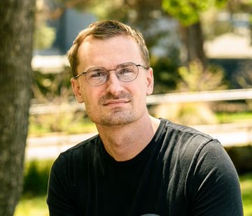 The image features a man wearing glasses sitting under a tree with his head tilted back. The background includes trees, suggesting that he might be outdoors or in a park setting. There are also some texts visible in the image, including one that reads "@base launches Base MCP", which could possibly be related to the post about AI agents controlling crypto wallets and token swapping on @coinbase's Ethereum L2.