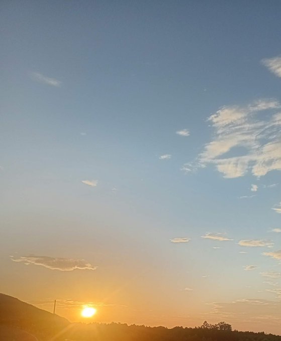 A scenic landscape photograph captures a bright orange sunset on the horizon beneath a vast blue sky scattered with wispy clouds.