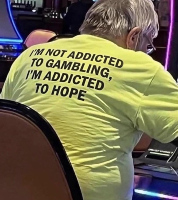 A person from behind wearing a yellow shirt with the text 'I'M NOT ADDICTED TO GAMBLING, I'M ADDICTED TO HOPE', sitting in front of slot machines.