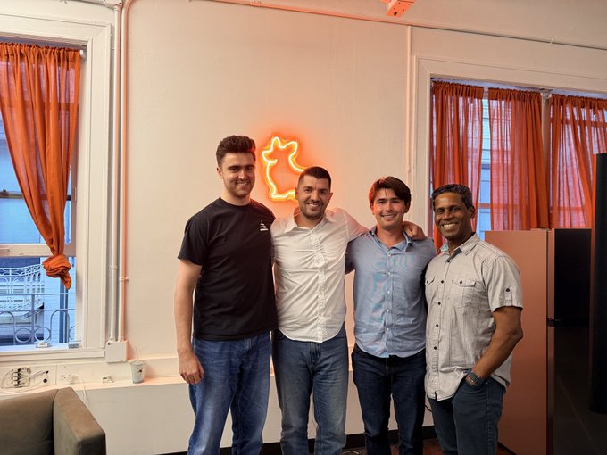 A photograph of four smiling men posing together indoors in front of an orange neon rabbit/fox sign, with orange curtains and windows visible in the background.