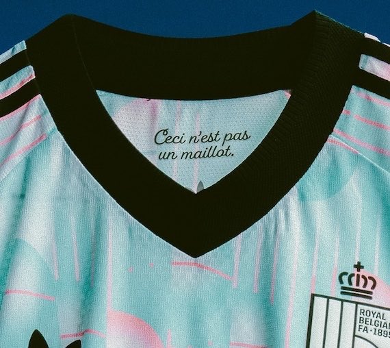 Close-up of Belgium's away soccer jersey featuring a black V-neck collar and a "Royal Belgian FA 1895" crest, with the interior label reading "Ceci n'est pas un maillot" in reference to René Magritte's iconic "The Treachery of Images." The pastel-colored design pays homage to the surrealist Belgian artist, blending football culture with fine art.