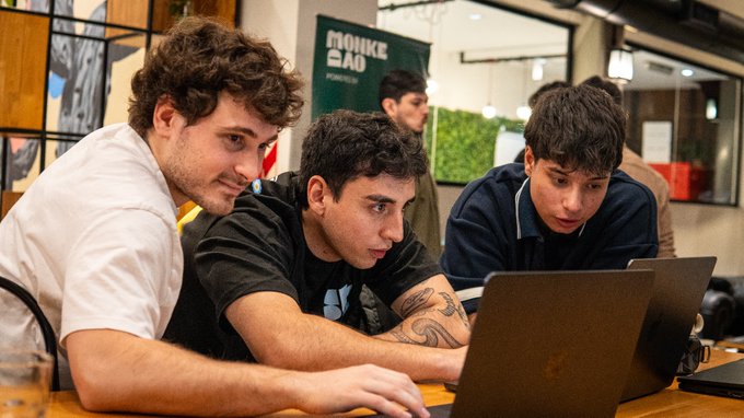 Three young men looking intently at a laptop screen at what appears to be a Monke salad restaurant, with one typing on the keyboard.