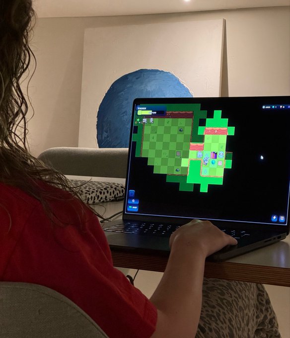 The image shows a person sitting at a desk, using a laptop computer with a game displayed on its screen. The game appears to be a strategy-based game called @onchainheroes MoG, which has been described as a "game changer" in terms of gameplay and mechanics. The player is wearing a red shirt while playing the game, adding a pop of color to the scene. The person's focus on the game suggests that they are engaged in an immersive gaming experience.