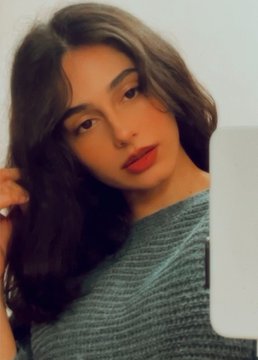 A young woman with long dark hair and red lipstick posing for a selfie while holding a white phone or object.