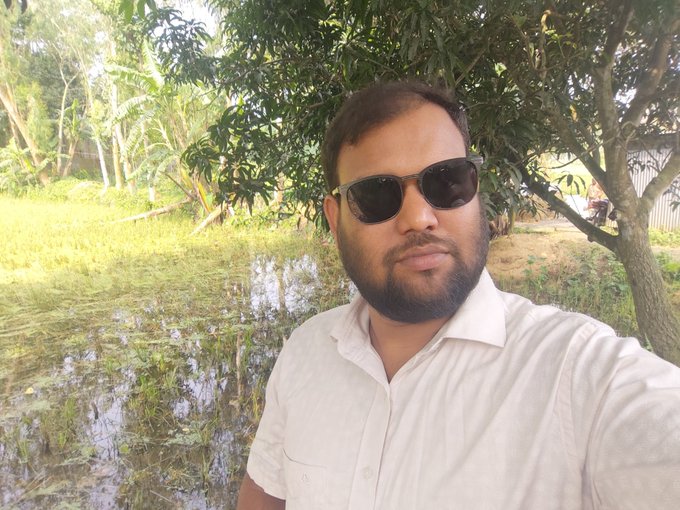 The image features a man wearing sunglasses and a white shirt, standing next to a tree with water around him. The background includes trees and bushes, creating an outdoor setting. There are also other people visible in the scene, but they are not the main focus of the photo. The man is looking directly at the camera, giving off a confident and relaxed vibe.