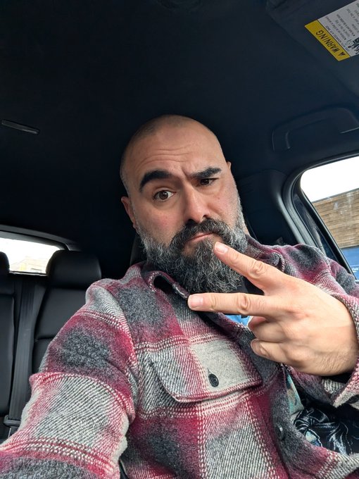 A bald man with a graying beard wearing a red and gray plaid flannel shirt takes a selfie inside a car, making a peace sign with his hand.