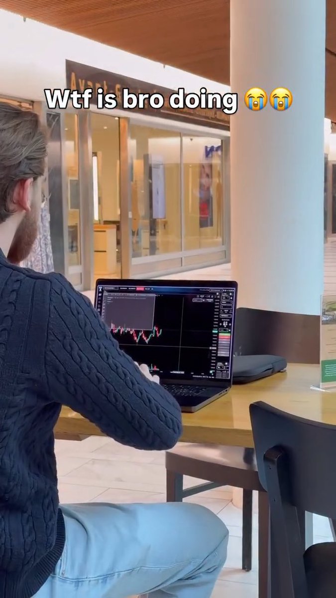 A man sits at a table in a mall using a laptop displaying stock trading charts, overlaid with the text "Wtf is bro doing 😭😭".