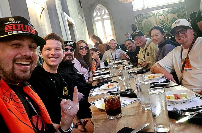 A large group of people smile and gesture around a long restaurant table during the ApeFest Builders Brunch. Visible text includes “ApeFest” and “RHINO” on a cap, “GAME” on another cap, plus ApeFest branding on clothing and lanyards.
