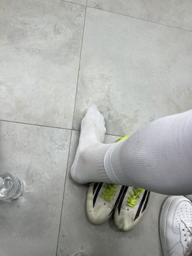 A person's feet in white socks standing on a tiled floor next to white and black sneakers, with a glass of water visible in the corner.