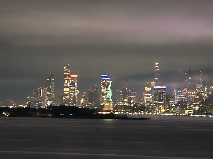 The Statue of Liberty stands illuminated against the glittering Manhattan skyline at night, with a bridge visible to the left—capturing the iconic image of New York City as a symbol of freedom and opportunity, tying into the post's message about pursuing massive financial success in America's financial capital.