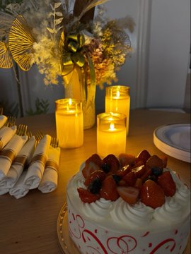A birthday or celebration table setting featuring a strawberry-topped cream cake, lit candles, rolled napkins with gold utensils, and a floral centerpiece with yellow flowers.