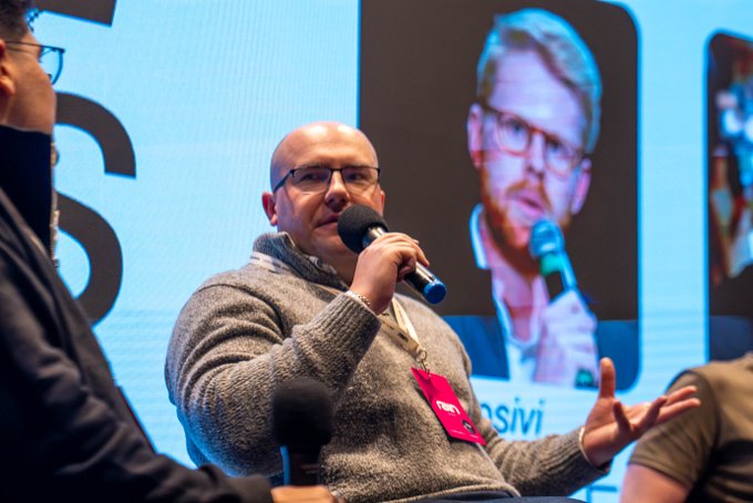 A panelist speaks into a microphone at the LATAM Digital Assets Conference, with the partial text "siv" visible in the background. A red "BUIDL" badge is attached to his gray sweater, and the screen behind displays another speaker, fitting the discussion on DeFi protocols featuring Olaf Kudin (1inch), Daniel Luca (OpenZeppelin), and nicnode (OctantApp).