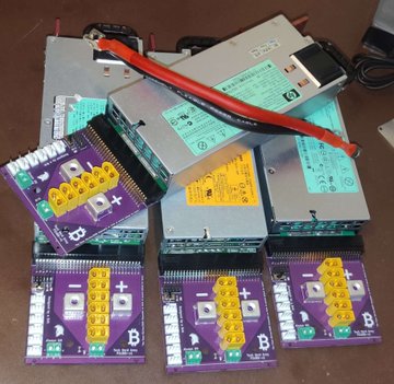 A pile of cryptocurrency mining power supplies and ASIC miner components, with one unit displaying a yellow connection board labeled 'D-', '+', 'B', and 'V'.