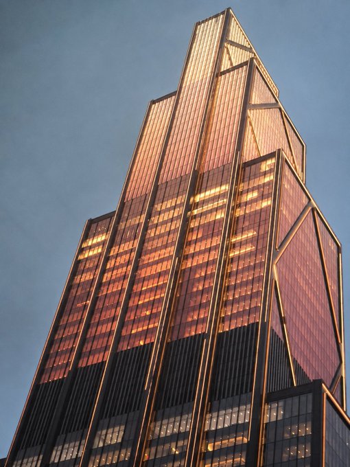 A low-angle photograph of a tall, modern glass skyscraper with a distinctive copper/bronze-colored geometric facade reflecting the sunlight against a clear blue sky.