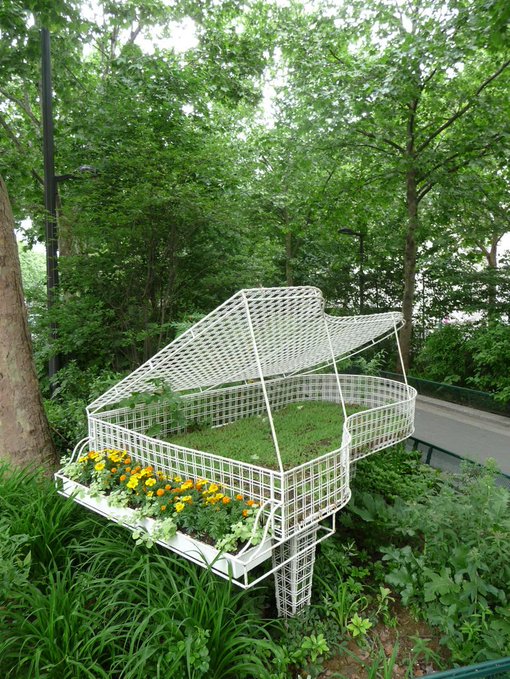 A creative outdoor garden installation featuring a white wire sculpture in the shape of a grand piano, filled with grass, yellow flowers, and greenery, set in a lush park area.