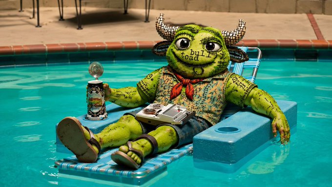 A green 'bulli' toad figurine with horns labeled 'bulli toad' on its face, lounging on a pool float in a swimming pool while holding a can and wearing a patterned shirt and sandals.