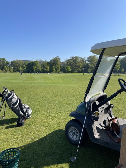 A golf cart with clubs parked beside a putting green under a clear blue sky, evoking the spirit of driving with passion as fellow golfers practice in the distance.