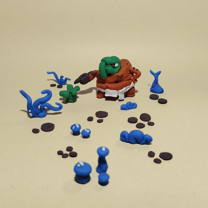 Claymation figures featuring a green-headed character in a karate stance surrounded by various small blue and green clay creations and pebbles.