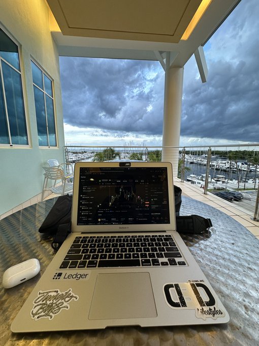 A sticker-covered silver laptop sits open on a textured outdoor table, displaying what appears to be a financial trading chart on its screen. The setup includes an AirPods case and overlooks a marina under a cloudy sky.