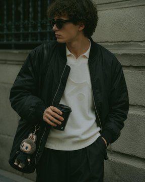 A man with curly hair and sunglasses poses in a street style look, wearing a black bomber jacket over a cream polo shirt, dark pants, and carrying a black crossbody bag with a small plush keychain. Holding a coffee cup, his aesthetic aligns with the "crypto Twitter" vibe referenced in the post—blending minimalist streetwear with subtle accessories suited for casual urban settings.