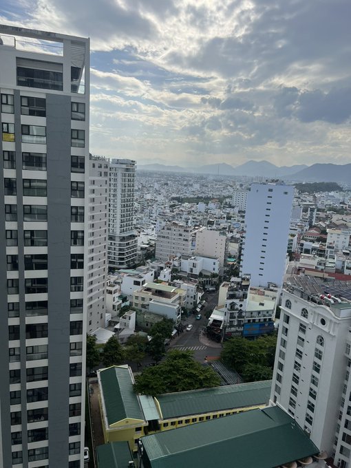 An elevated cityscape view shows high-rise apartment buildings, streets with traffic, and a mountain range in the distance under a partly cloudy sky.