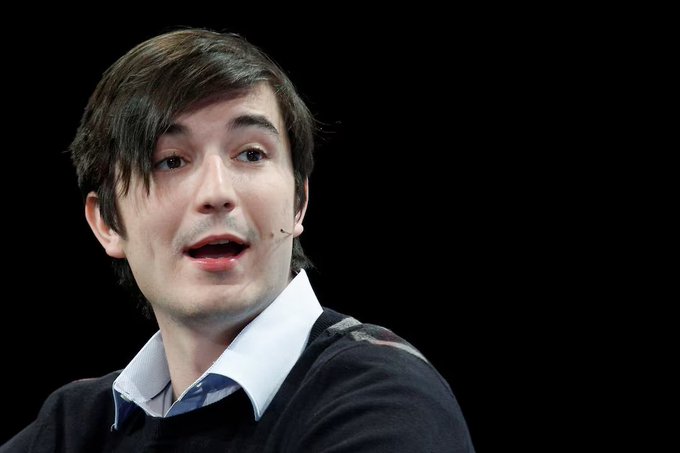A man with dark hair wearing a light blue collared shirt and dark sweater speaks while wearing a small earpiece microphone against a black background.