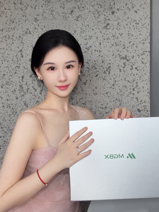 A young woman in a pink lace dress smiles while holding a white laptop displaying the green 'M2EX' logo against a textured gray wall.