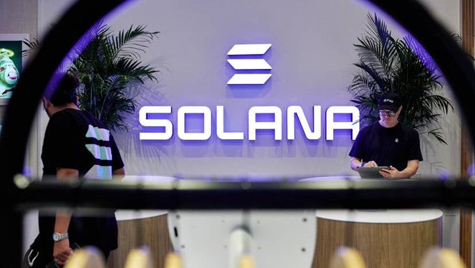 A person in a black cap stands at a reception desk in front of a large illuminated "SOLANA" logo flanked by tropical plants, while another person walks past carrying a bag. The image visually represents the cryptocurrency brand mentioned in the post about Solana securing a default position for stablecoins at an FDIC-insured bank.