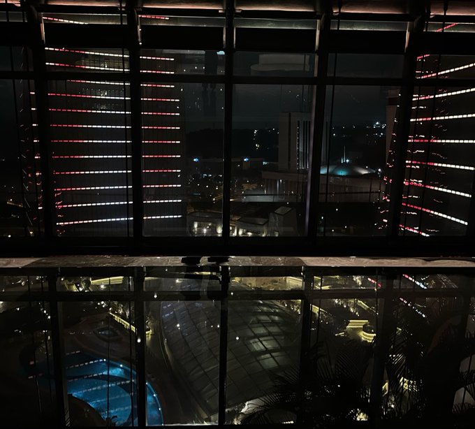 A nighttime view through large grid-framed windows showing illuminated buildings with red and white lights, with reflections of a pool and indoor space visible in the glass below.