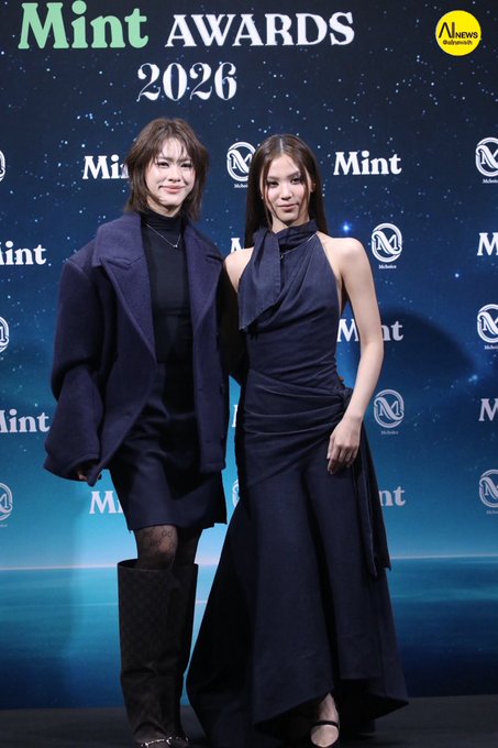 Two women posing on a red carpet at the 'Mint AWARDS 2026' event, one wearing an oversized dark coat and knee-high boots, the other in a navy halter-neck gown.
