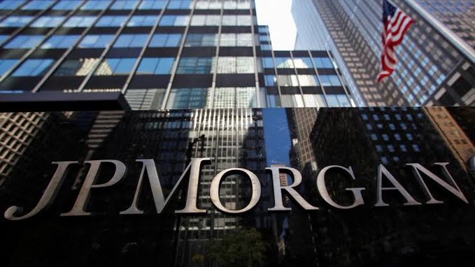 The JPMorgan logo is displayed on a dark reflective surface, with the bank's modern glass skyscraper and an American flag visible in the background.