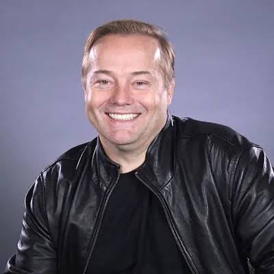 A confident man with short brown hair and a wide smile, wearing a black leather jacket over a black t-shirt, poses for a portrait against a muted gray background. The image accompanies a bearish opinion on Bitcoin's price recovery, suggesting skepticism about its near-term prospects despite the recent rebound.
