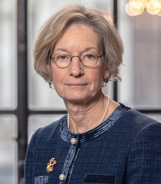 A portrait photo shows an older woman with short gray hair, glasses, a blue textured jacket with a gold brooch, and a pearl necklace.