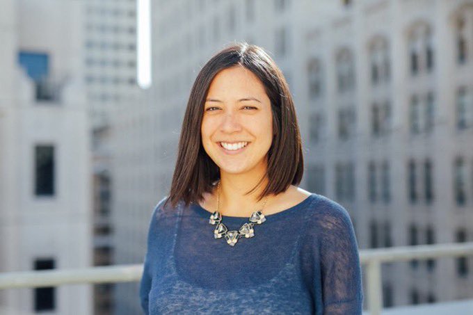 A professional headshot of a smiling woman with shoulder-length dark hair, wearing a blue top and a statement necklace, posed against a blurred urban background of tall buildings.