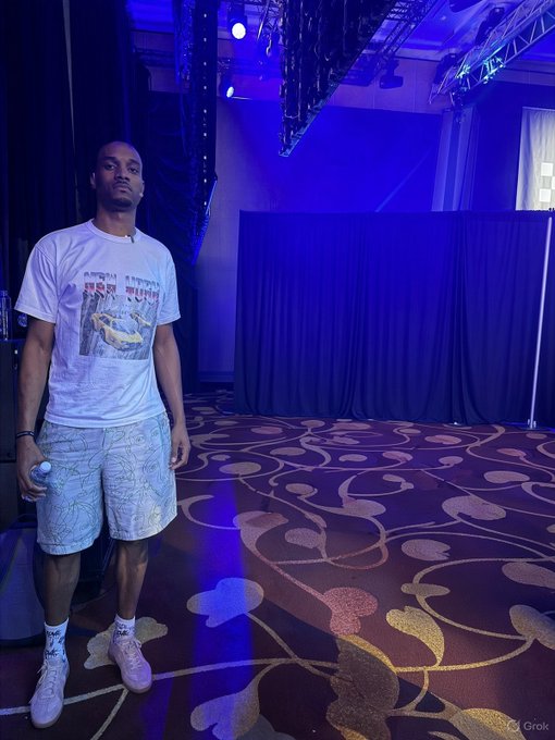 A man in a white graphic t-shirt and patterned shorts standing in a dimly lit room with blue stage lighting and patterned carpet, likely a concert or event venue.
