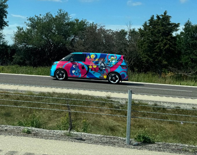 A colorful car with cartoon character graphics drives along a rural road bordered by trees and a wire fence.