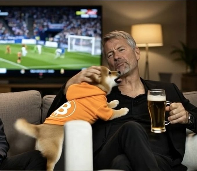 An older man with gray hair and beard sitting on a couch holding a beer mug while watching a soccer match on TV, as a Shiba Inu dog wearing an orange hoodie stands on its hind legs interacting with him.